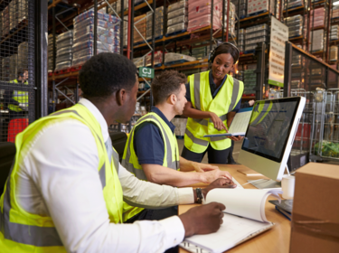 Three people in safety vests at a computer in a warehouse